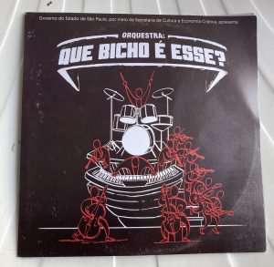 Fotografia da capa do libreto da orquestra Que Bicho é Esse?. Fundo preto e o desenho de uma bateria ao centro.
