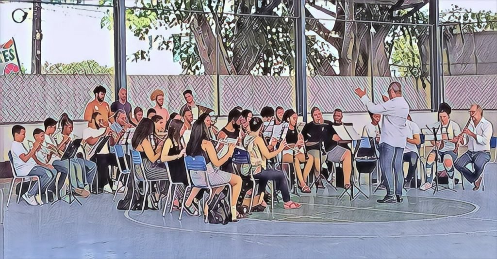 Pessoas sentadas e com instrumentos musicais, em aula com um maestro em uma quadra escolar. Por Todas as Bandas do Brasil abre inscrições