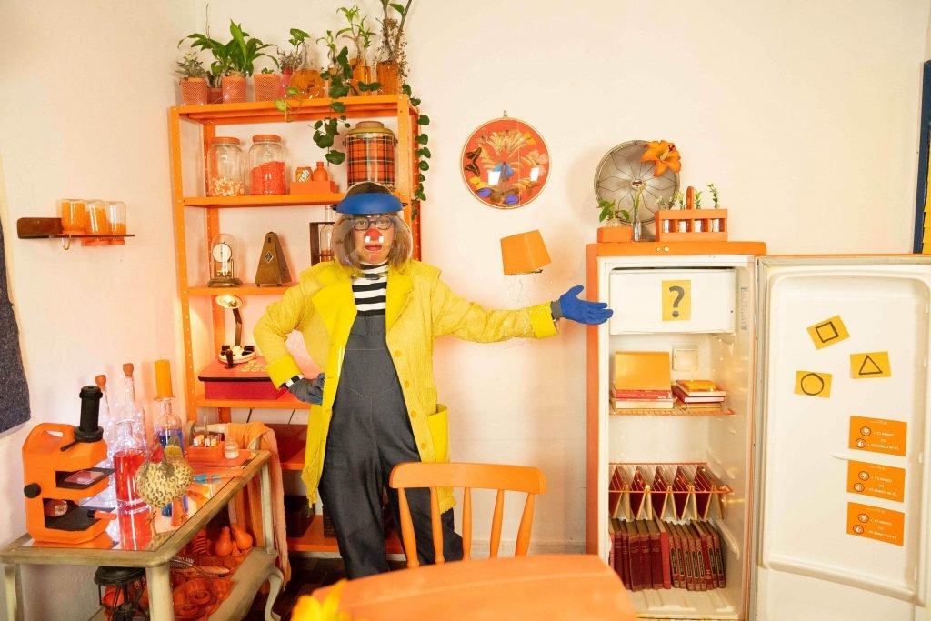 Sala com mesa, geladeira e estante laranjas. Uma mulher palhaça com capa amarela.